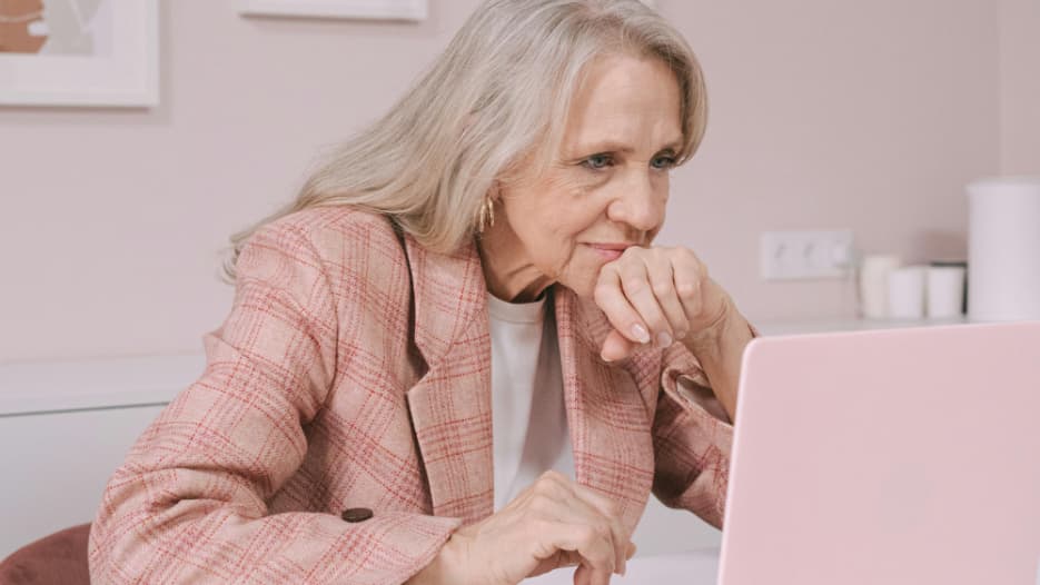 Ältere Dame sitzt am Schreibtisch und vergleicht Festgeldangebote verschiedener Banken auf einem Laptop