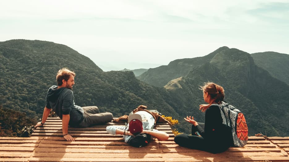 Drei Wanderer entspannen auf einer Holzplattform mit Blick auf eine beeindruckende Berglandschaft.