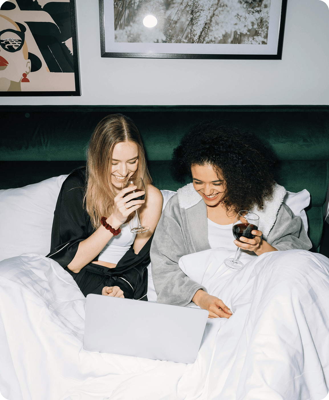 Zwei Frauen lachen entspannt im Bett mit Laptop und Weingläsern